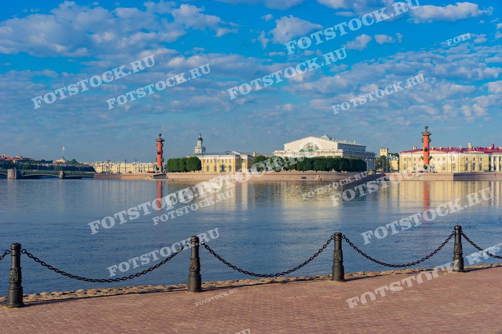 На фотографии изображён вид на Стрелку Васильевского острова в Санкт-Петербурге. Центральное место занимает здание Биржи и две Ростральные колонны, которые отражаются в воде Невы. Снимок сделан в летний день, что подчёркивается ярким солнцем и голубым небом. На переднем плане видна часть набережной с цепями и фонарями, добавляющая кадру глубины.
